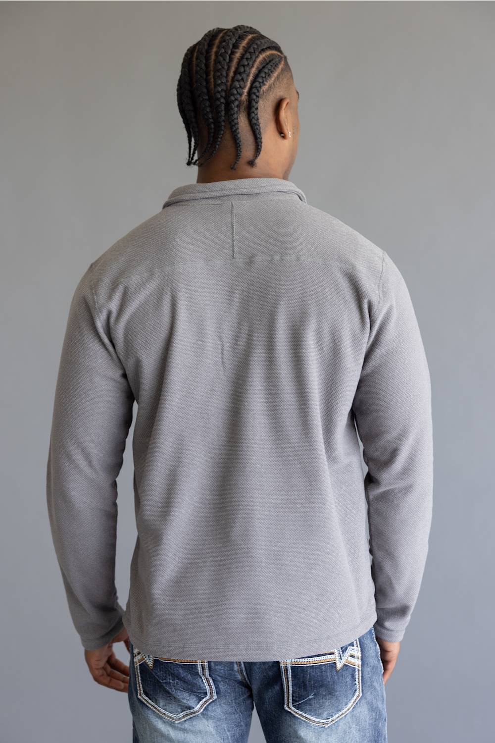 A man viewed from behind wearing a light gray long-sleeve knit polo shirt and blue jeans, standing against a plain gray studio background.