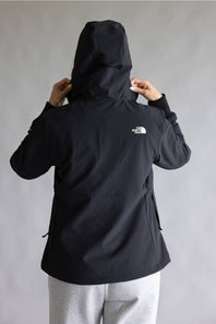 A person shown from the back wearing a black hooded jacket with a small white The North Face logo on the upper back, paired with light gray sweatpants, against a plain gray studio background.