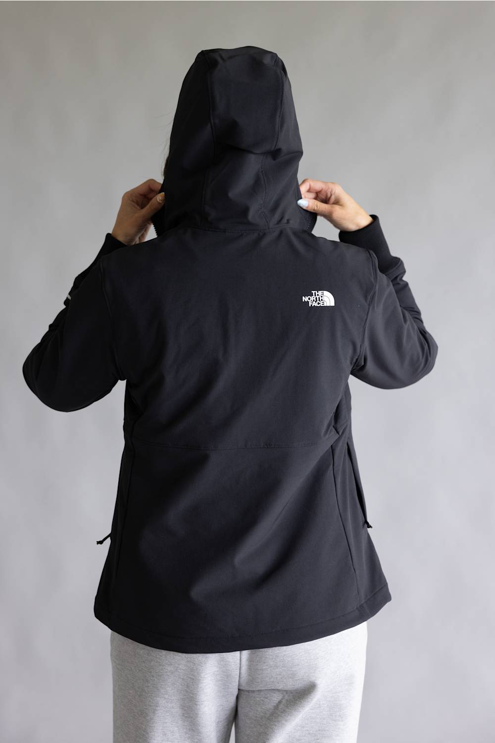 A person shown from the back wearing a black hooded jacket with a small white The North Face logo on the upper back, paired with light gray sweatpants, against a plain gray studio background.