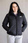 A woman wearing a black quilted puffer jacket by The North Face, paired with light gray sweatpants, standing against a plain light gray background.