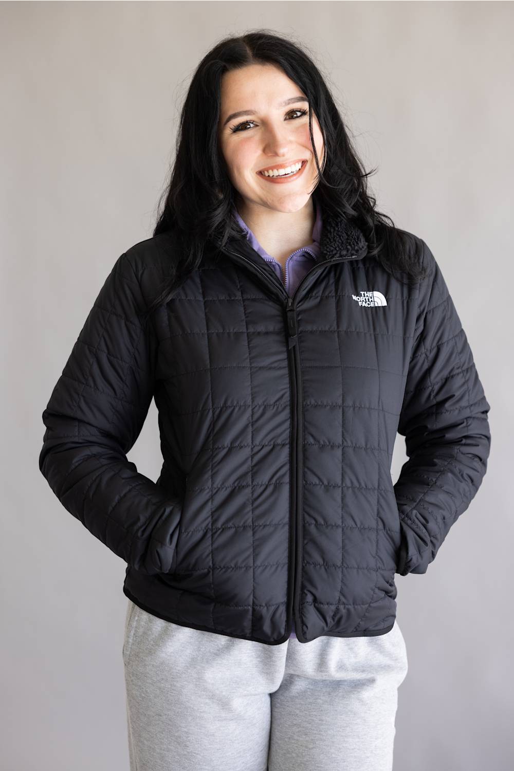 A woman wearing a black quilted puffer jacket by The North Face, paired with light gray sweatpants, standing against a plain light gray background.