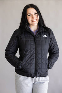 A woman wearing a black quilted puffer jacket by The North Face, paired with light gray sweatpants, standing against a plain light gray background.