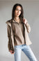A woman modeling a beige quarter-zip pullover sweater layered under a light brown zip-up hooded jacket, paired with light-wash blue jeans, against a solid gray studio background.