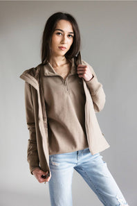 A woman modeling a beige quarter-zip pullover sweater layered under a light brown zip-up hooded jacket, paired with light-wash blue jeans, against a solid gray studio background.