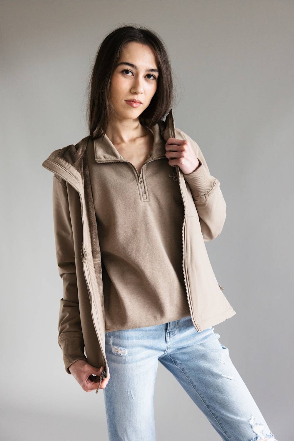 A woman modeling a beige quarter-zip pullover sweater layered under a light brown zip-up hooded jacket, paired with light-wash blue jeans, against a solid gray studio background.