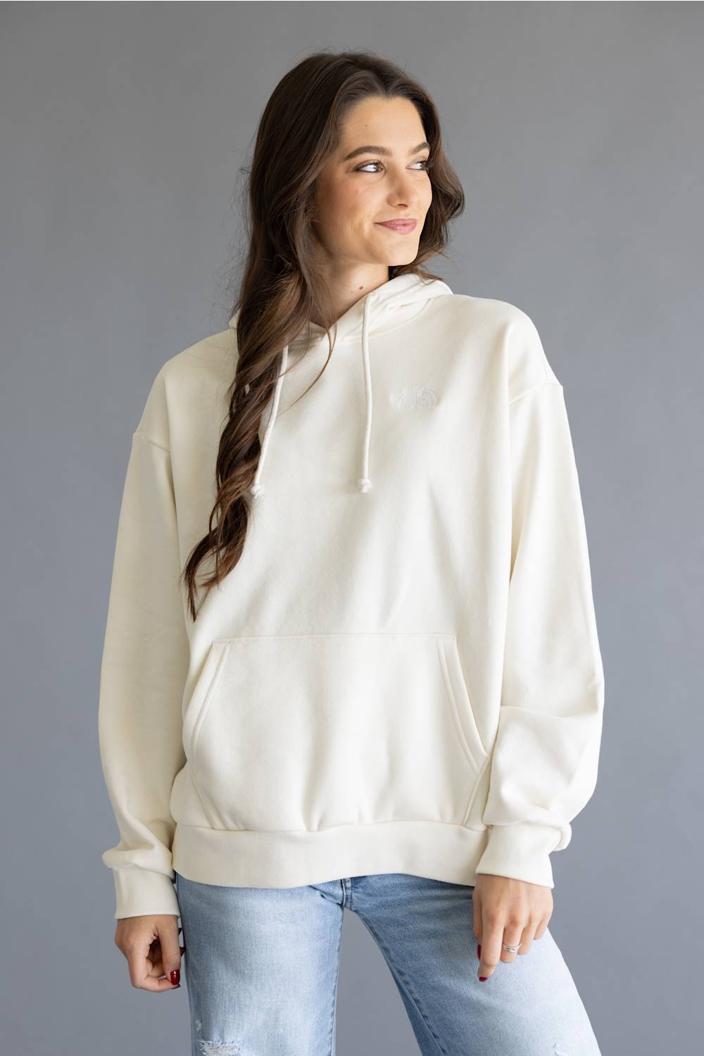 A woman wearing an oversized cream-colored pullover hoodie with drawstrings and a kangaroo pocket, paired with light blue jeans, standing against a solid gray studio background.