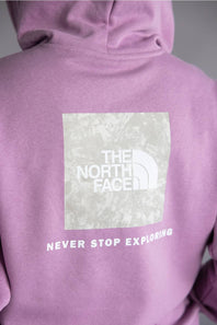 Close-up of the back of a lavender-purple hoodie featuring a large white square logo patch for The North Face with the text 'NEVER STOP EXPLORING' printed below it.