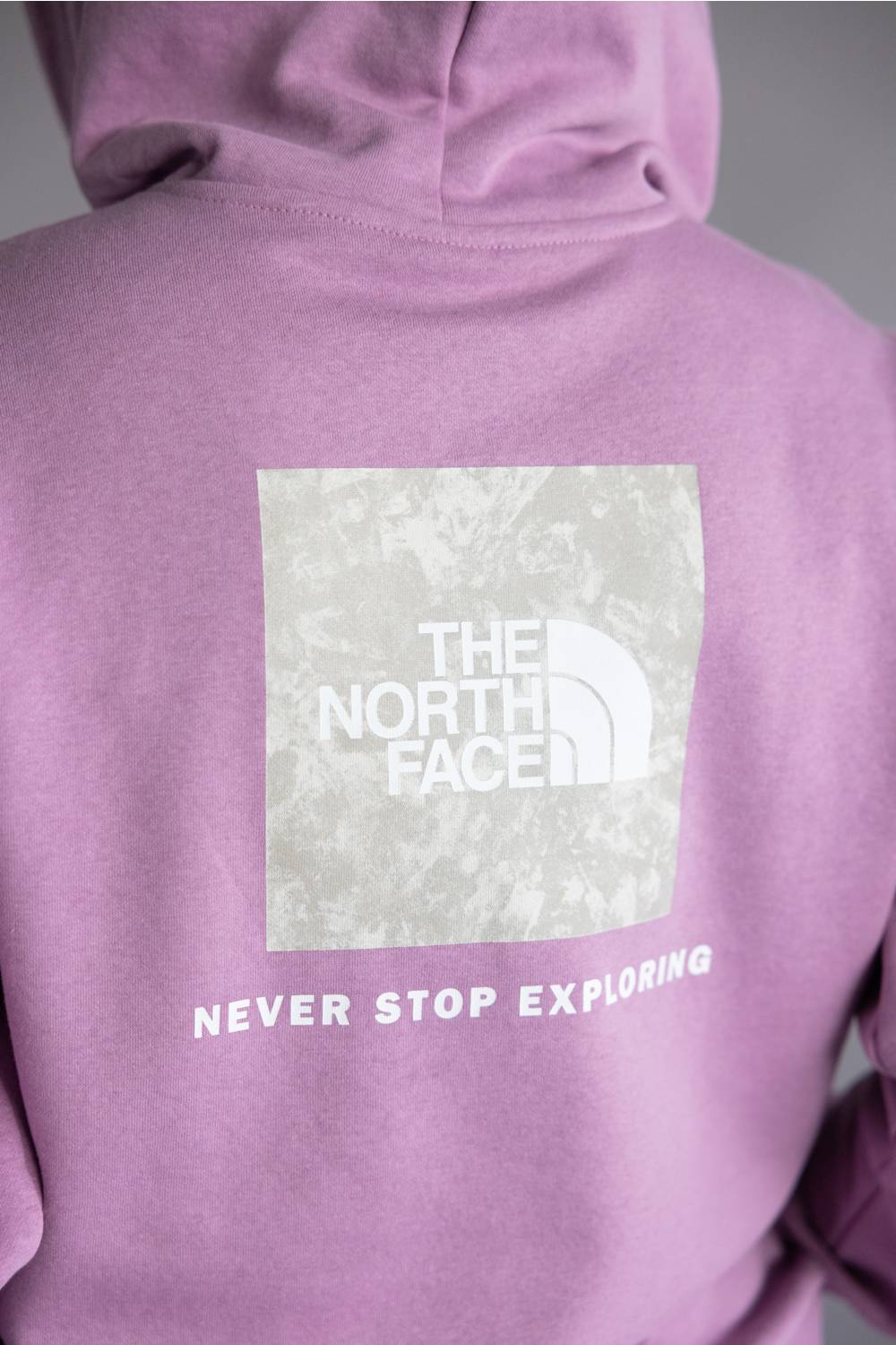 Close-up of the back of a lavender-purple hoodie featuring a large white square logo patch for The North Face with the text 'NEVER STOP EXPLORING' printed below it.