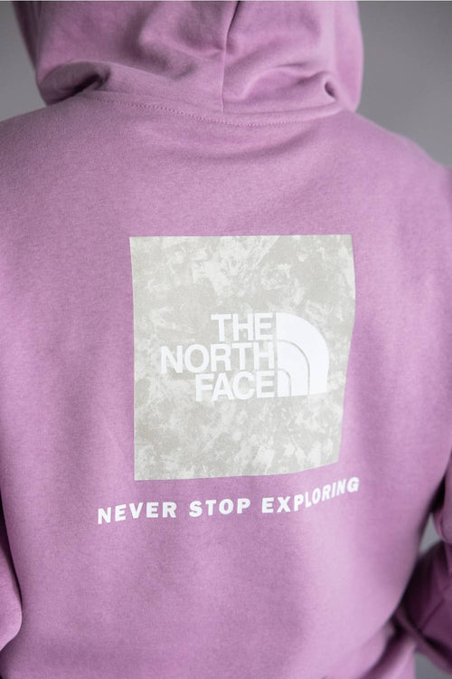 Close-up of the back of a lavender-purple hoodie featuring a large white square logo patch for The North Face with the text 'NEVER STOP EXPLORING' printed below it.
