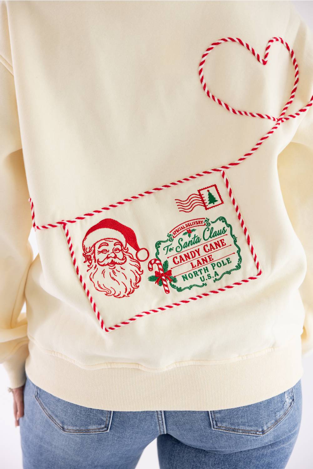 A close-up of a cream-colored sweatshirt featuring a red and green Christmas-themed envelope graphic with Santa Claus, a candy cane, and a heart outline, worn by a model with blue jeans.