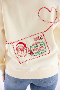 A close-up of a cream-colored sweatshirt featuring a red and green Christmas-themed envelope graphic with Santa Claus, a candy cane, and a heart outline, worn by a model with blue jeans.
