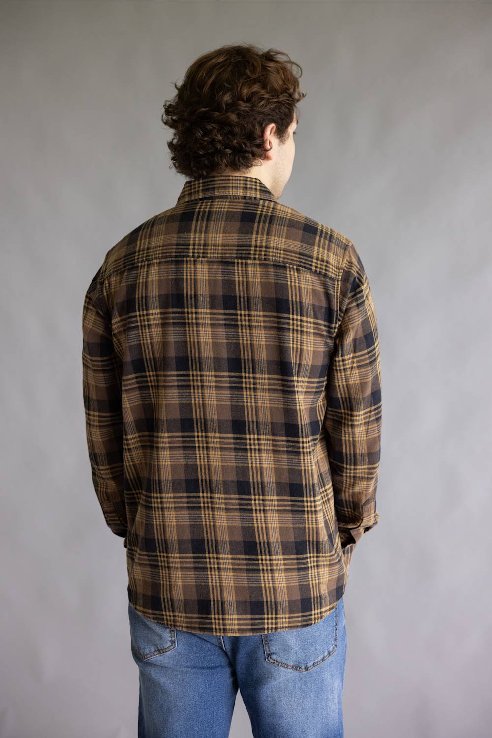 A man viewed from behind wearing a brown and black plaid flannel shirt with blue jeans, standing against a neutral gray studio background.
