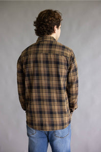 A man viewed from behind wearing a brown and black plaid flannel shirt with blue jeans, standing against a neutral gray studio background.