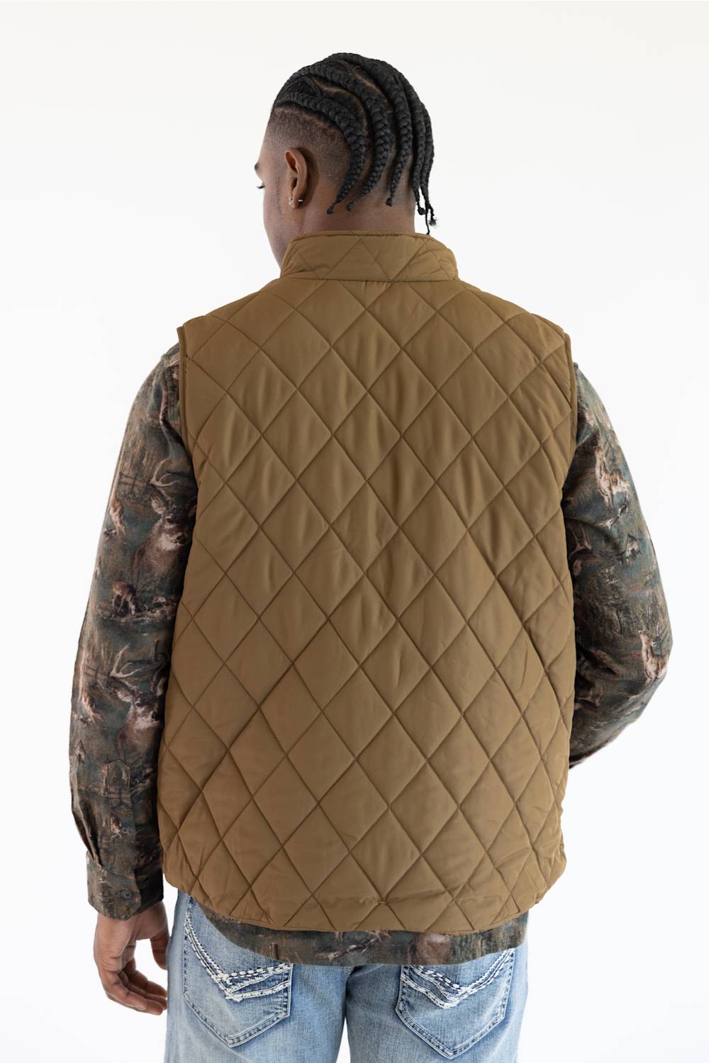 A man viewed from the back wearing a tan diamond-quilted puffer vest over a camouflage long-sleeve shirt and blue jeans, against a plain white background.
