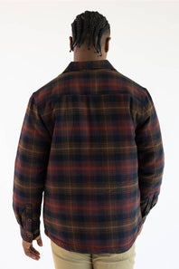 A man viewed from behind wearing a long-sleeve plaid button-up shirt in dark navy, rust red, and tan, paired with khaki pants, against a plain white background.