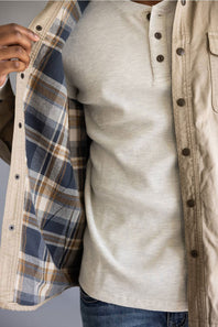 A close-up of a person wearing a light beige ribbed henley shirt under an open tan overshirt with a blue, beige, and brown plaid lining.