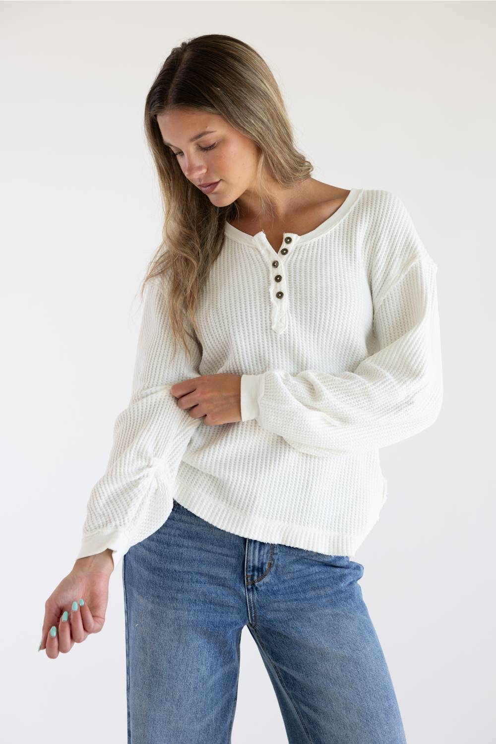 A woman wearing a white ribbed long-sleeve henley sweater with button details and blue jeans, standing against a plain white background.