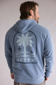 Back view of a person wearing a blue hoodie with a palm tree design and 'Jetty' text on the back. The hoodie is Jetty Orbit Grove Hoodie for Men in Pigment Slate