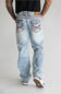 Rear view of a person wearing light-wash blue jeans with heavily embellished multicolor embroidered back pockets, paired with a black shirt and white sneakers, against a light gray studio background.