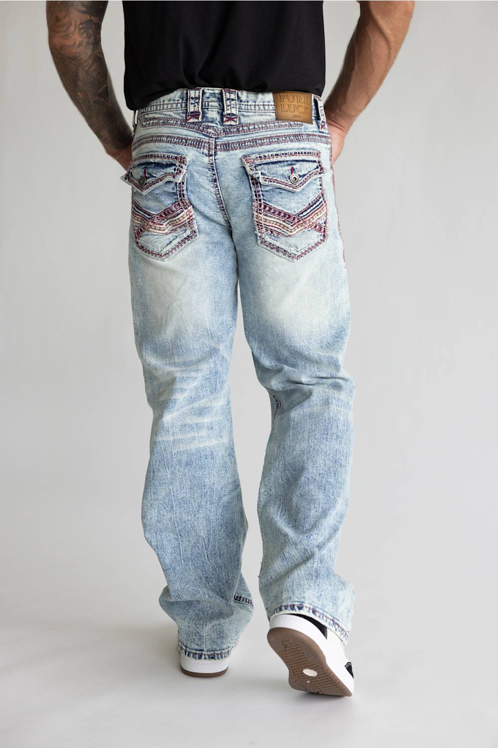 Rear view of a person wearing light-wash blue jeans with heavily embellished multicolor embroidered back pockets, paired with a black shirt and white sneakers, against a light gray studio background.