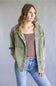 A woman wearing an olive green corduroy button-up shirt jacket over a brown tank top and light blue jeans, standing against a neutral gray studio background.