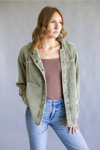 A woman wearing an olive green corduroy button-up shirt jacket over a brown tank top and light blue jeans, standing against a neutral gray studio background.