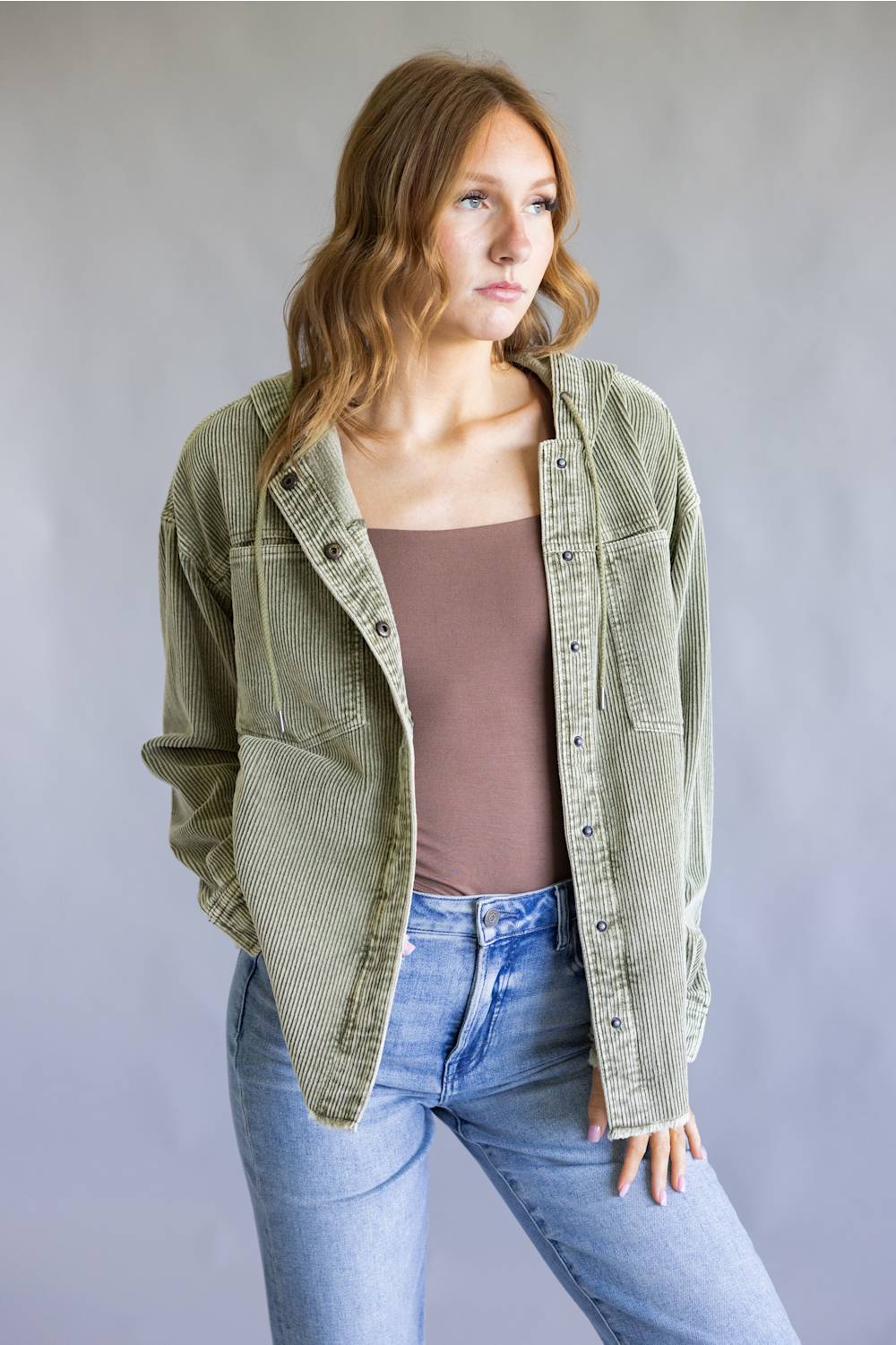 A woman wearing an olive green corduroy button-up shirt jacket over a brown tank top and light blue jeans, standing against a neutral gray studio background.
