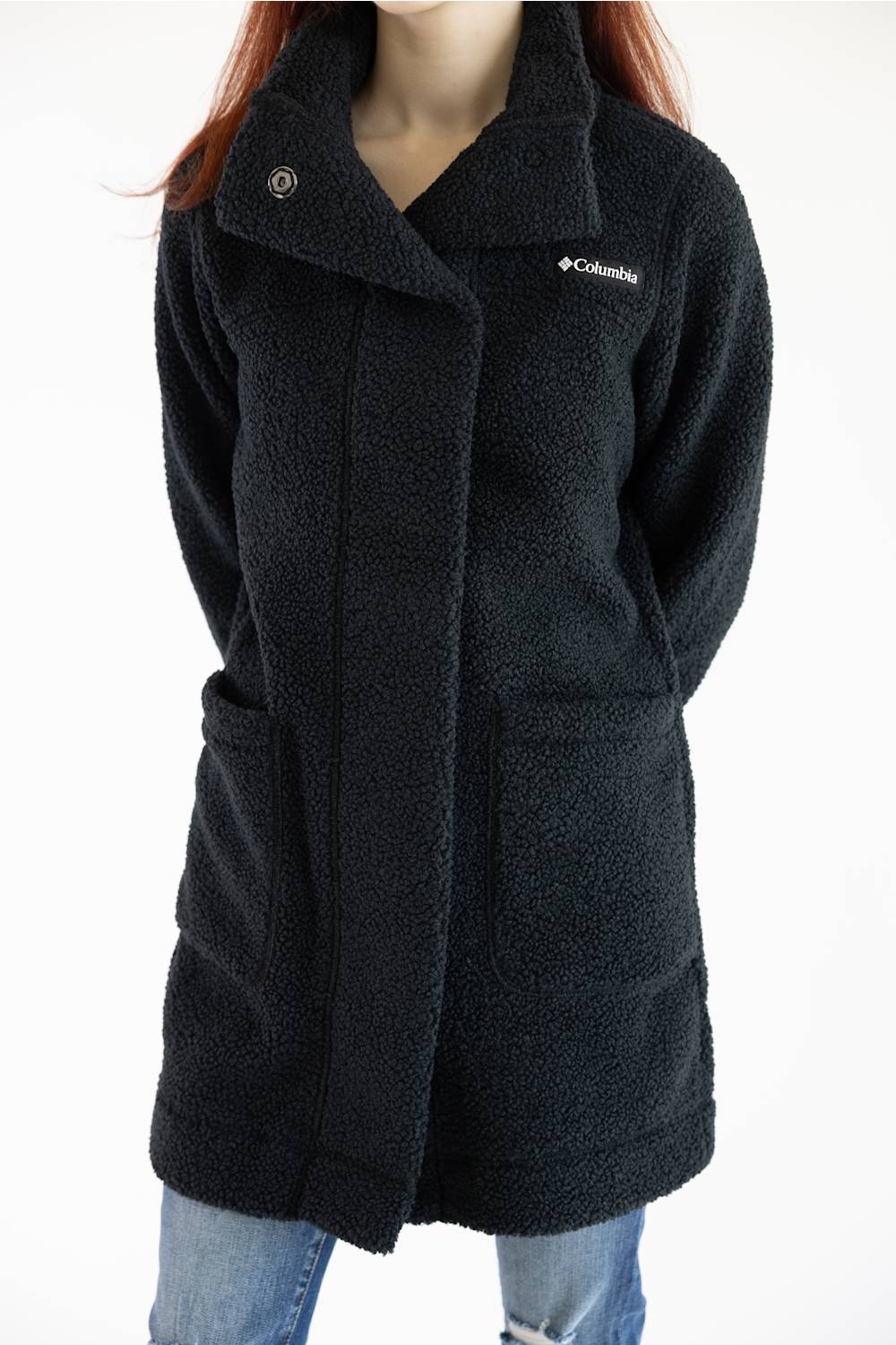 A woman wearing a black Columbia sherpa fleece coat with a snap-button closure, large front patch pockets, and a small white Columbia logo on the chest, paired with blue jeans against a plain white background.