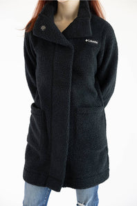 A woman wearing a black Columbia sherpa fleece coat with a snap-button closure, large front patch pockets, and a small white Columbia logo on the chest, paired with blue jeans against a plain white background.