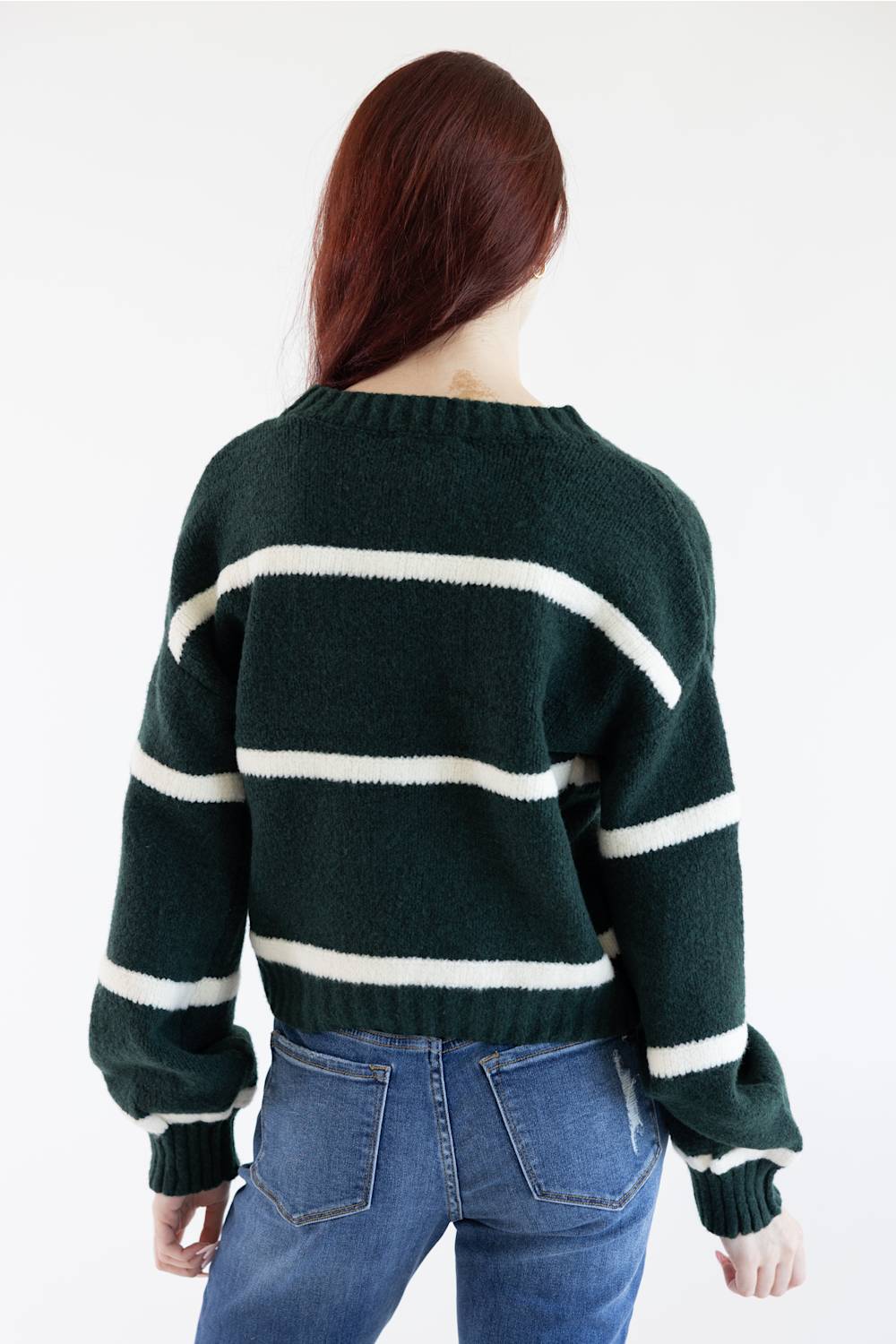 A woman viewed from behind wearing a dark green knit sweater with horizontal cream stripes and blue jeans, against a plain white studio background.