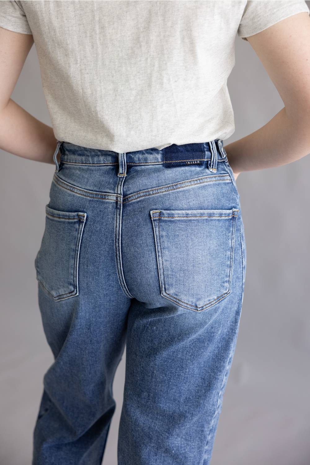 Close-up of a person's hands adjusting the waistband and button of blue denim jeans, wearing a light gray heathered t-shirt.