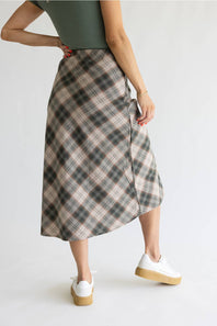 A woman wearing a mid-length plaid wrap skirt in beige, gray, and black, paired with a dark green top and white platform sneakers, shown from a side-back angle against a plain white studio background.