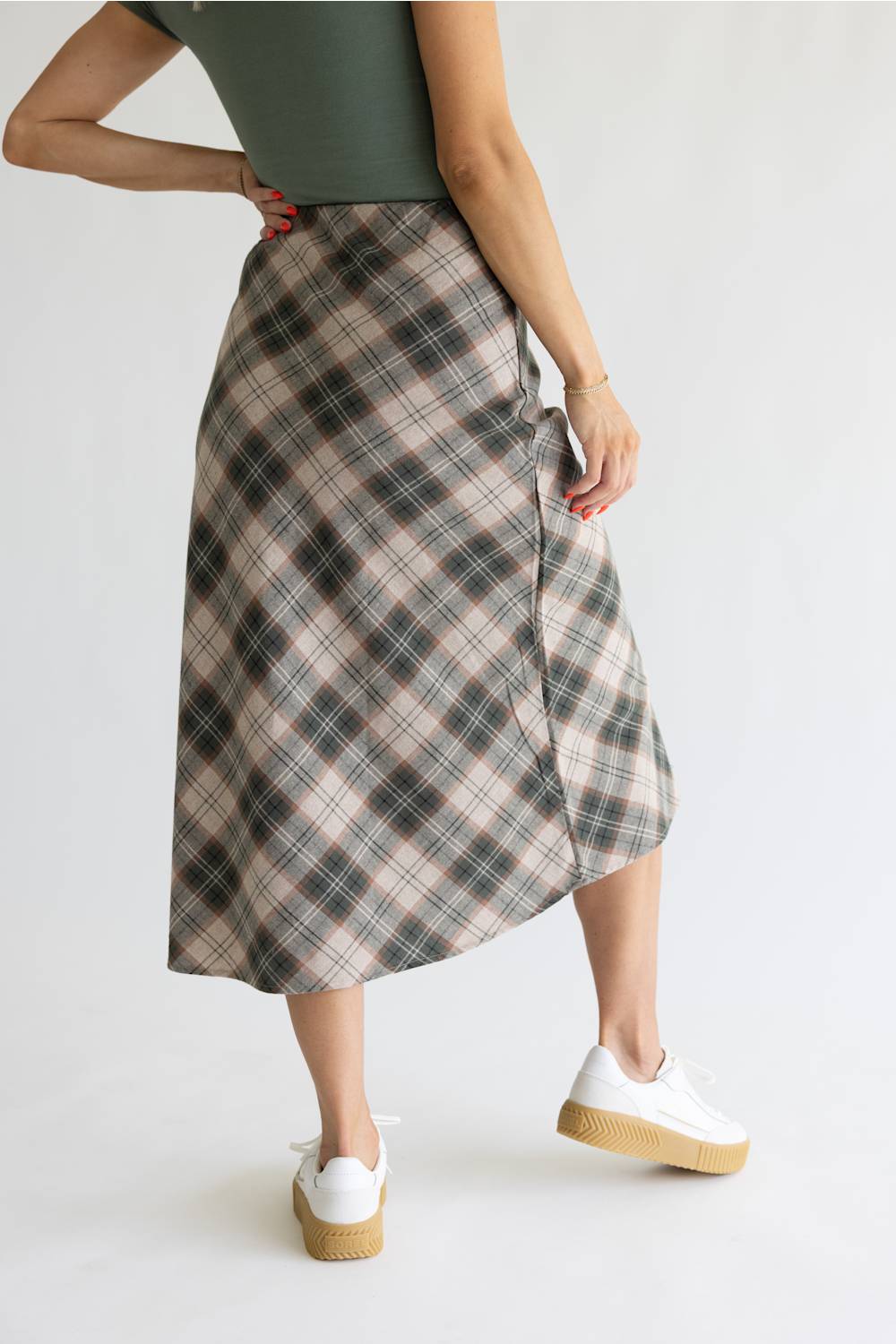 A woman wearing a mid-length plaid wrap skirt in beige, gray, and black, paired with a dark green top and white platform sneakers, shown from a side-back angle against a plain white studio background.