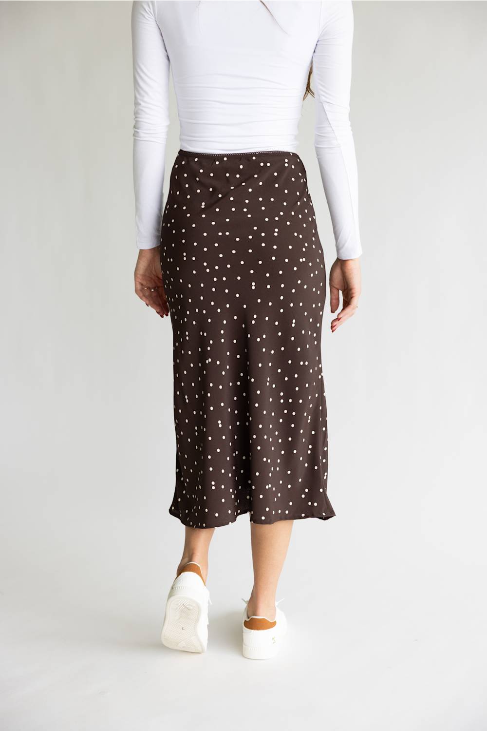 A model from the back wearing a brown midi skirt with a white polka dot pattern, styled with a white long-sleeve top and white platform sneakers against a light gray studio background.