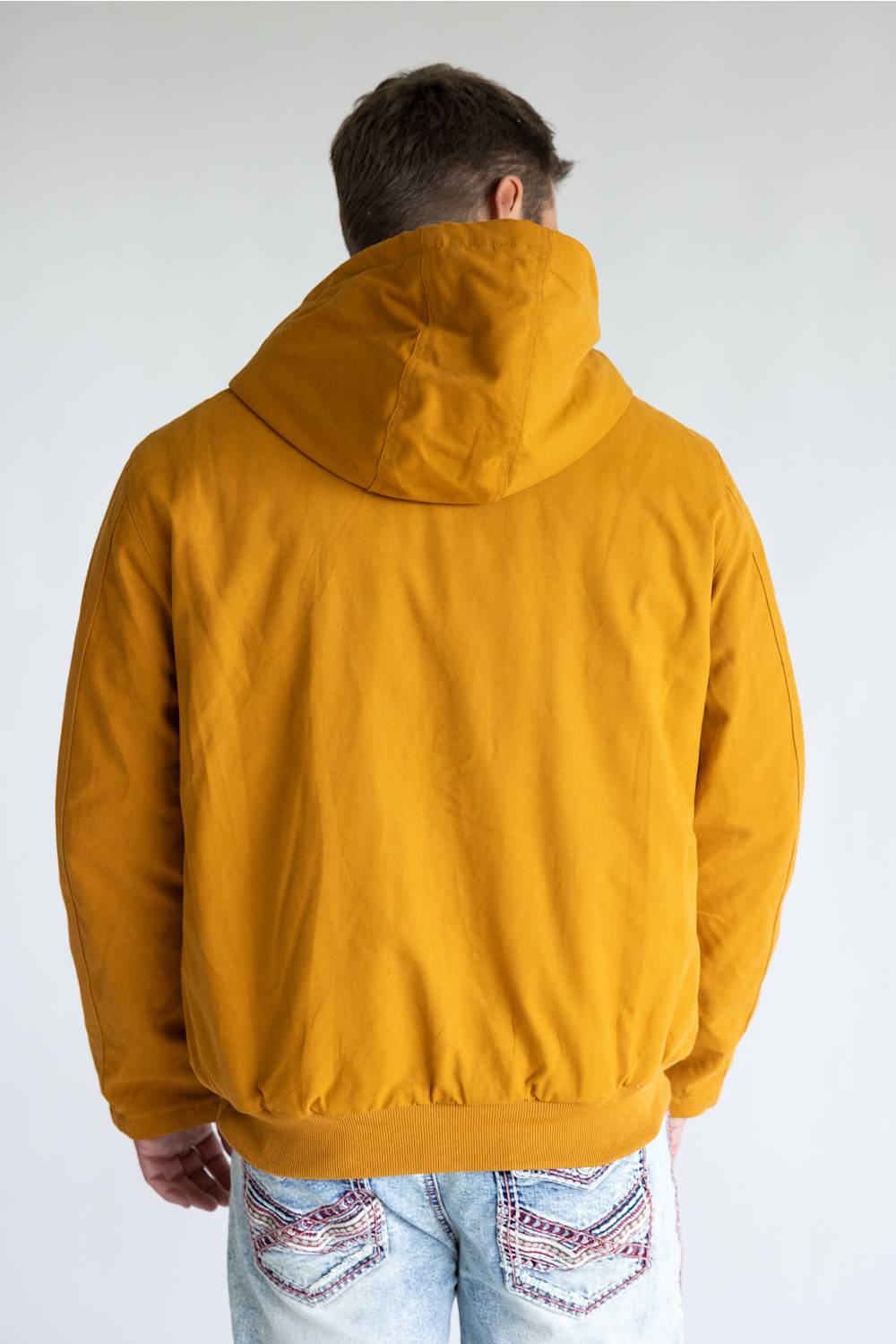 A man viewed from behind wearing a mustard yellow hooded jacket and light blue jeans with embroidered details, set against a plain light gray background.
