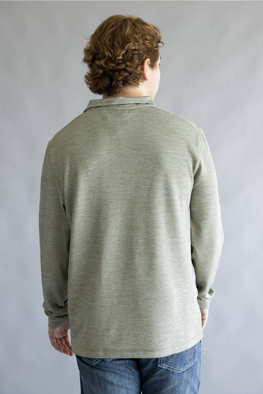 A man viewed from behind wearing a light sage green long-sleeve knit polo shirt and blue jeans, standing against a neutral gray studio background.