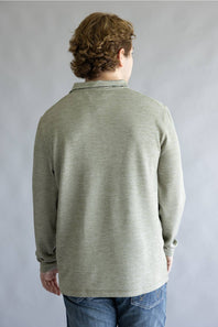 A man viewed from behind wearing a light sage green long-sleeve knit polo shirt and blue jeans, standing against a neutral gray studio background.