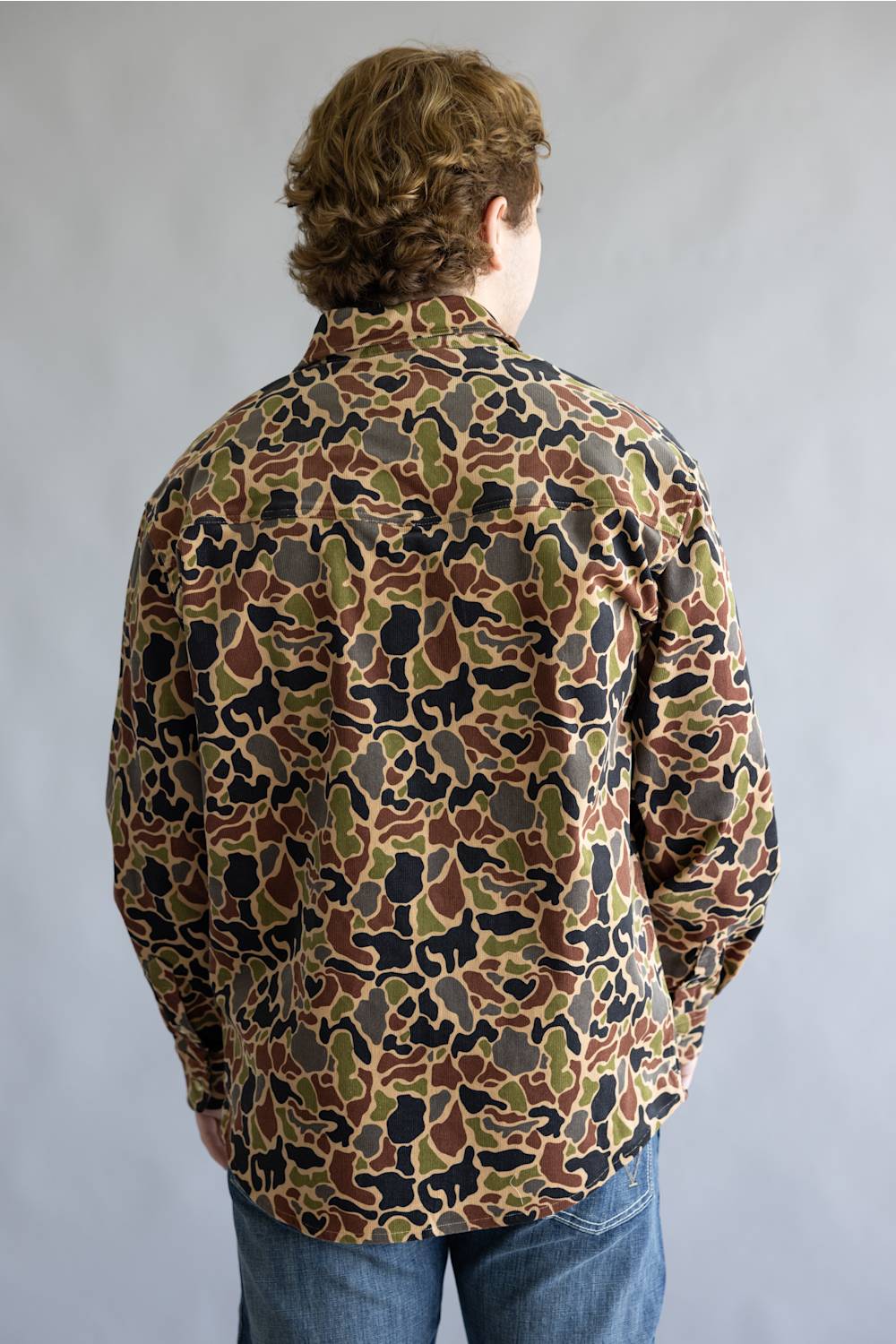 A person viewed from the back wearing a long-sleeve button-up shirt with an all-over camouflage pattern in brown, olive green, black, and tan, paired with blue jeans against a plain gray studio background.
