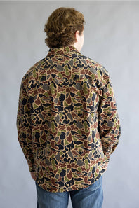 A person viewed from the back wearing a long-sleeve button-up shirt with an all-over camouflage pattern in brown, olive green, black, and tan, paired with blue jeans against a plain gray studio background.