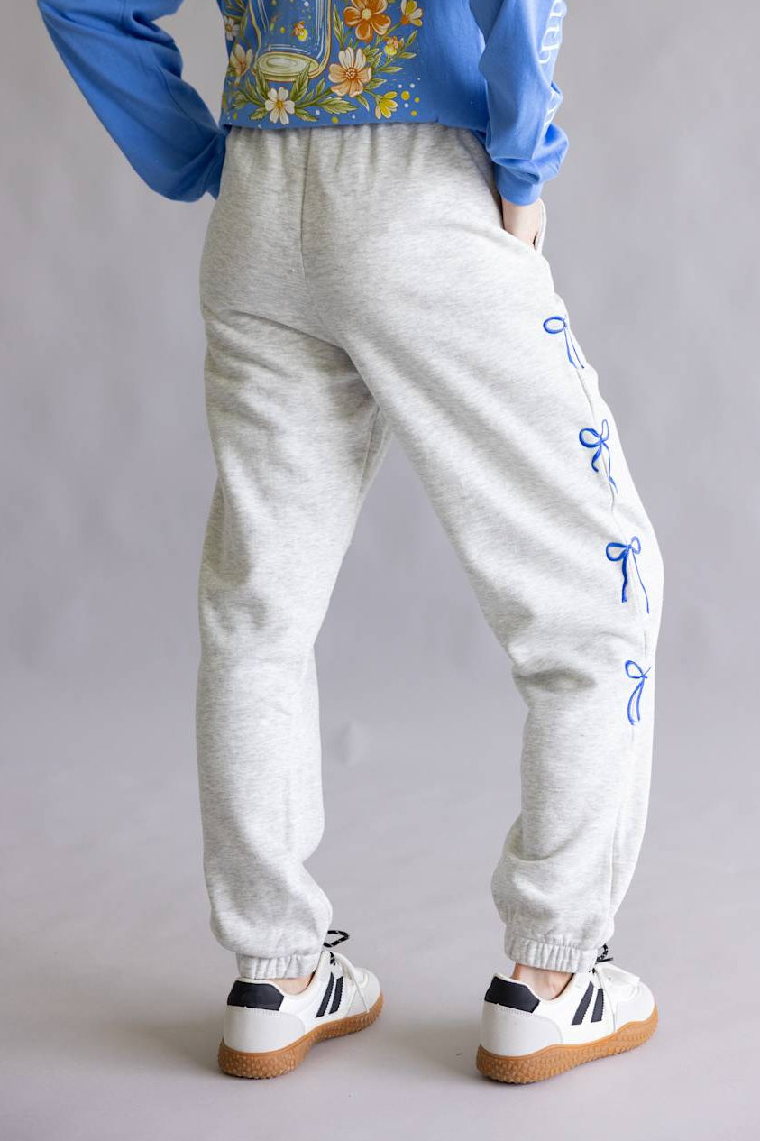 A person shown from the back wearing light heather gray jogger sweatpants with blue embroidered bow details down the sides, a blue long-sleeve top with a floral and jar graphic, and white sneakers with black stripes.