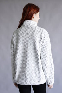 A woman with red hair is shown from the back wearing a white textured fleece pullover sweater with a mock neck, paired with black pants, against a plain gray studio background.