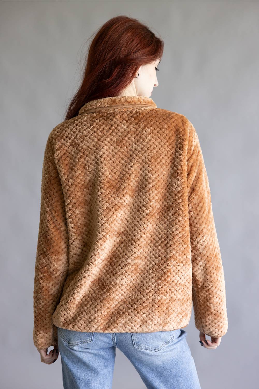 A woman with long red hair is shown from the back wearing a textured, warm tan-brown fuzzy jacket and light blue jeans against a plain gray studio background.