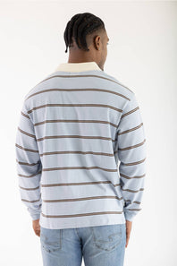 A man viewed from behind wearing a light blue long-sleeve striped shirt with brown horizontal stripes and a cream collar, paired with light blue jeans, against a plain white background.