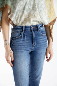 A close-up of a person wearing medium-wash blue denim jeans and a patterned cream blouse with brown trim, cropped from the chest to the upper thighs against a plain light background.