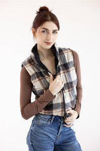 A woman wearing a brown long-sleeve top and blue jeans, featuring a beige, brown, and black plaid zip-up vest over her shoulders, set against a plain white studio background.