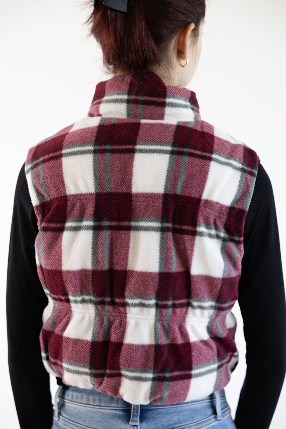 A person shown from the back wearing a sleeveless plaid fleece vest over a black long-sleeve shirt and blue jeans. The vest features a high collar and a burgundy, white, and gray plaid pattern.