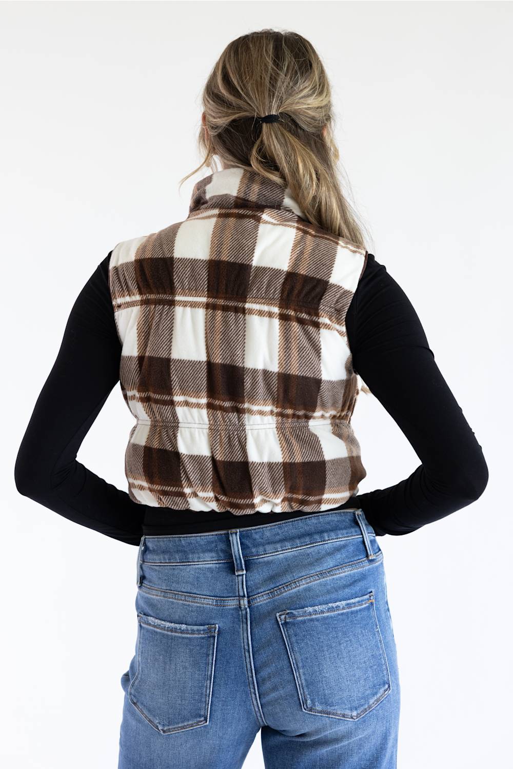 A woman viewed from behind wearing a brown and cream plaid sleeveless vest over a black long-sleeve top and blue jeans, against a plain white background.