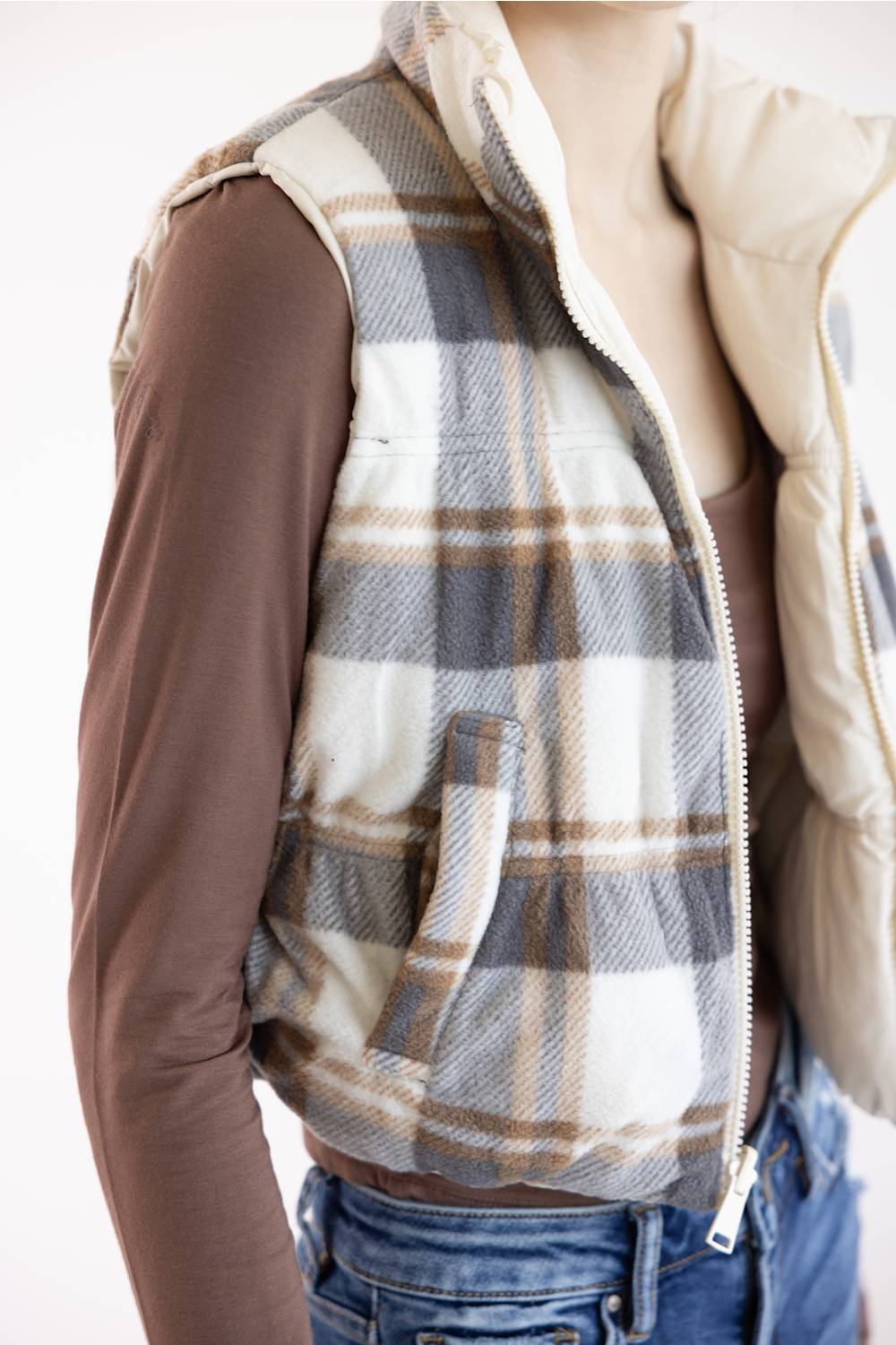 A sleeveless zip-front vest with a beige, gray, and brown plaid pattern, worn over a brown long-sleeve shirt and blue jeans.