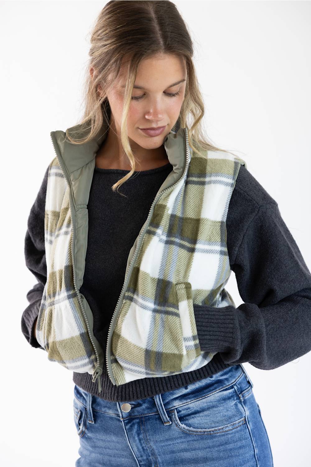 A woman wearing a cropped zip-front vest with a green, white, and beige plaid pattern over a dark charcoal knit sweater and blue jeans, photographed against a plain white studio background.