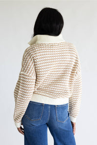 A person shown from the back wearing a cream and beige open-knit cropped sweater with a ribbed turtleneck and hem, paired with blue jeans, against a plain light gray studio background.